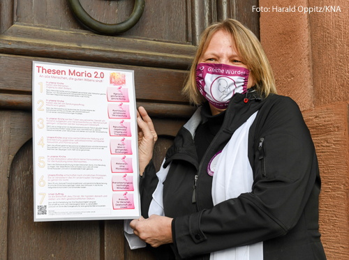 Andrea Keber hält ein Plakat in der Hand, dass sie gleich an eine Kirchentür schlagen wird. Darauf stehen sieben Thesen der Initiative Maria 2.0 für Reformen in der katholischen Kirche.