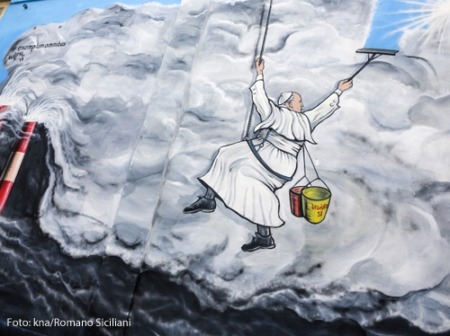 Ein Graffiti in Rom zeigt eine Figur von Papst Franziskus, der wie ein Fensterputzer an einem Seil hängt und Rauchwolken - Smog - wegwischt, so dass blauer Himmel und Sonne zum Vorschein kommen. Er hat einen Eimer dabei, auf dem "Laudato Si" steht. Links im Bild stehen rote Schornsteine, die die Umwelt verschmutzen.