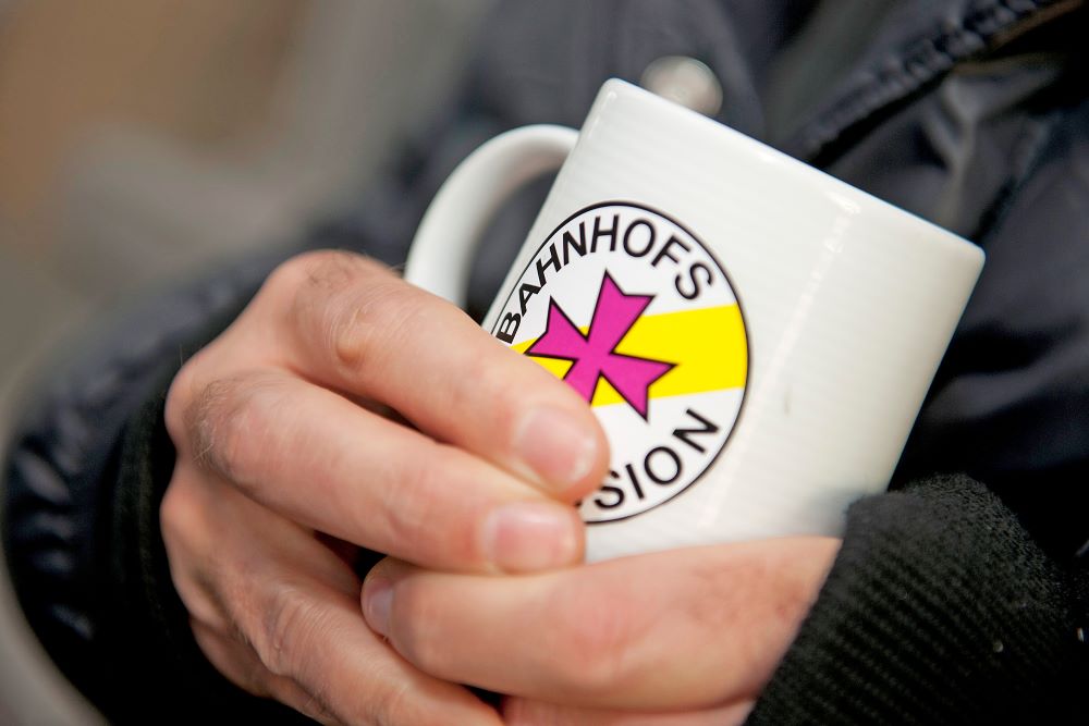 Jemand hält eine Tasse mit dem Logo der Bahnhofsmission in der Hand
