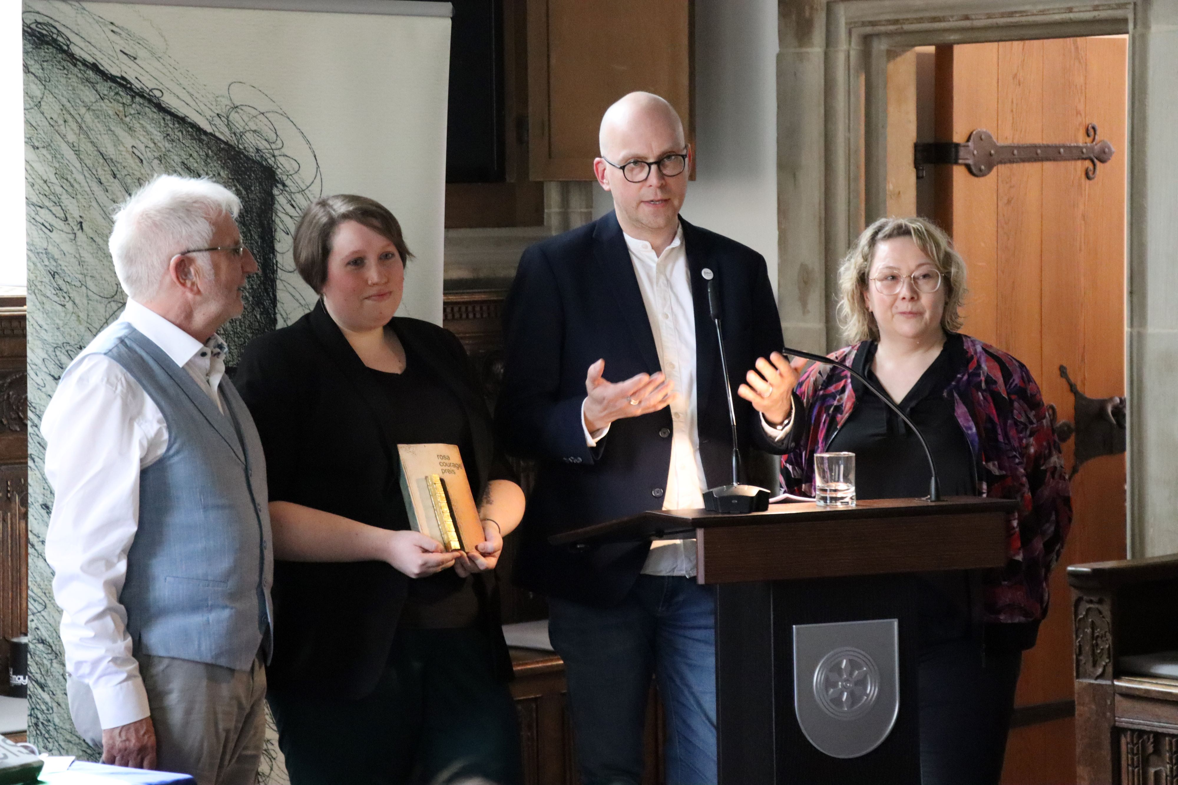 Ein Mann steht vor einem Rednerpult. Um ihn herum stehen zwei Frauen und ein Mann.