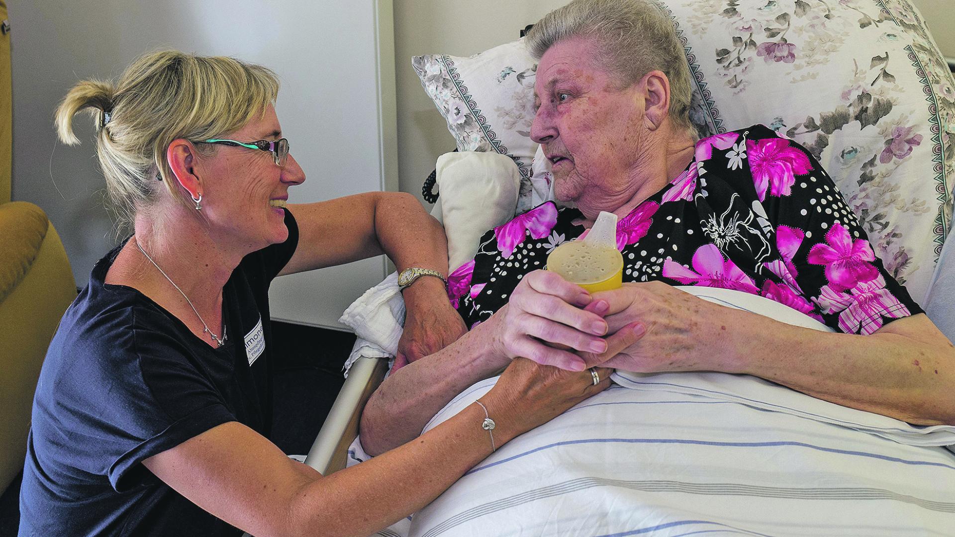 Eine Schwester reicht einer im Bett liegenden Person einen Trinkbecher.
