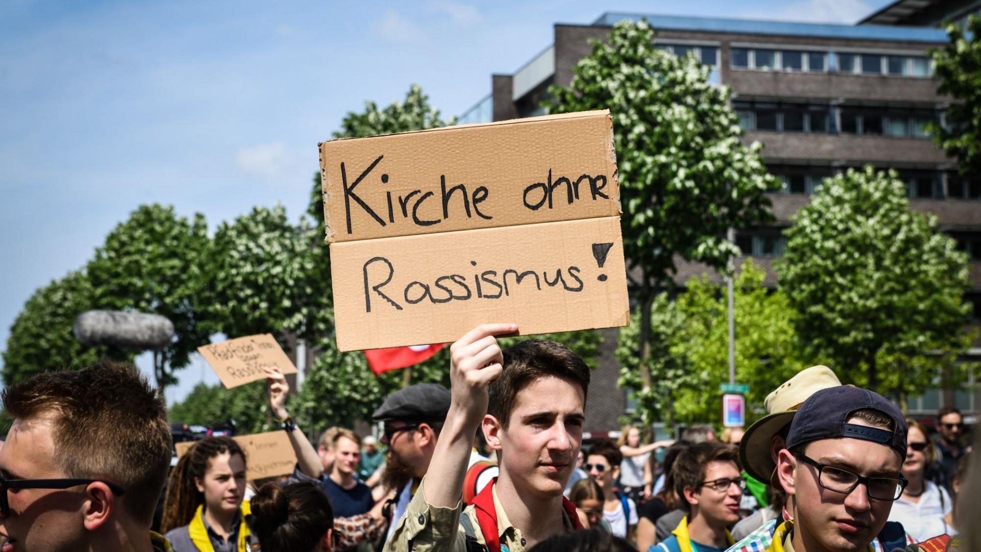 Ein Demonstrant hält bei einem Protest beim Katholikentag 2018 in Münster ein Schild mit der Aufschrift "Kirche ohne Rassismus" in die Höhe