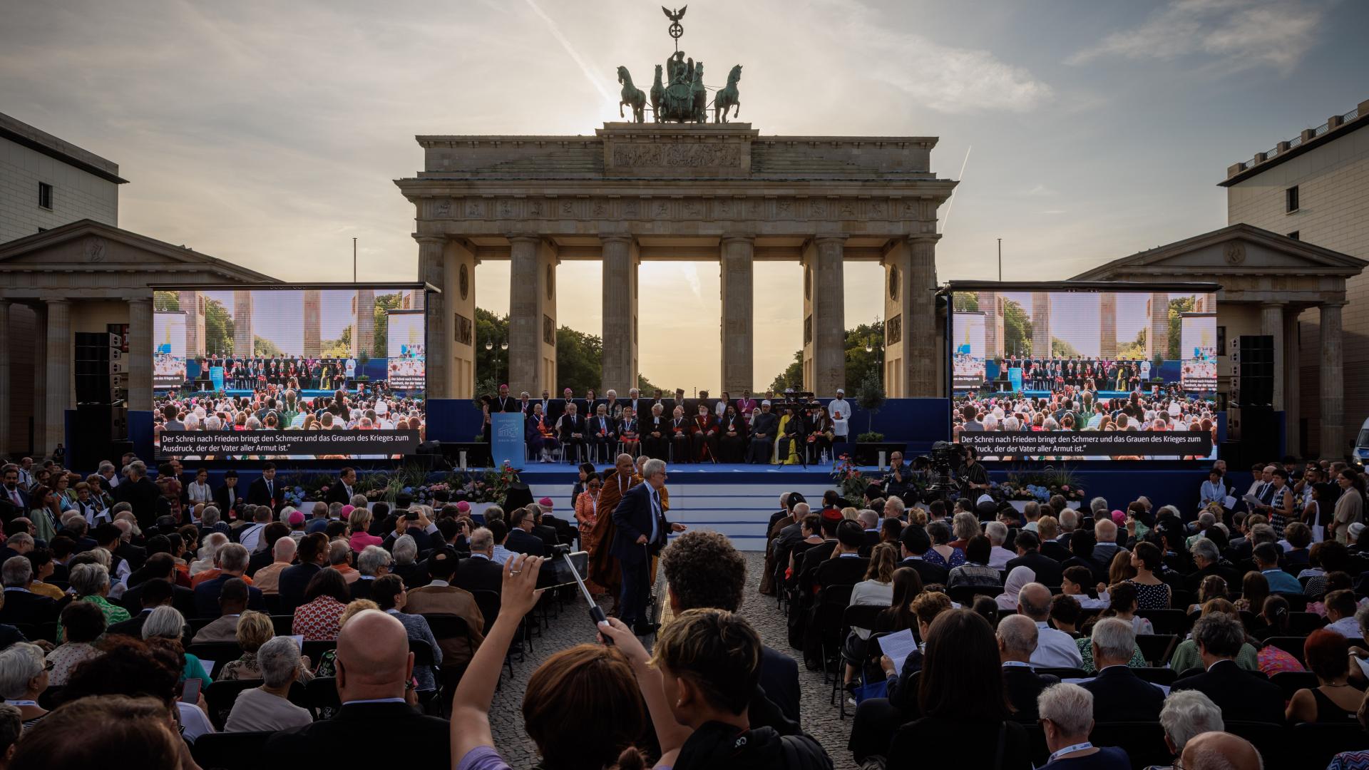 Foto: Menschen sitzen und stehen bei der Abschlussveranstaltung des Friedenstreffens St.Egidio in Berlin
