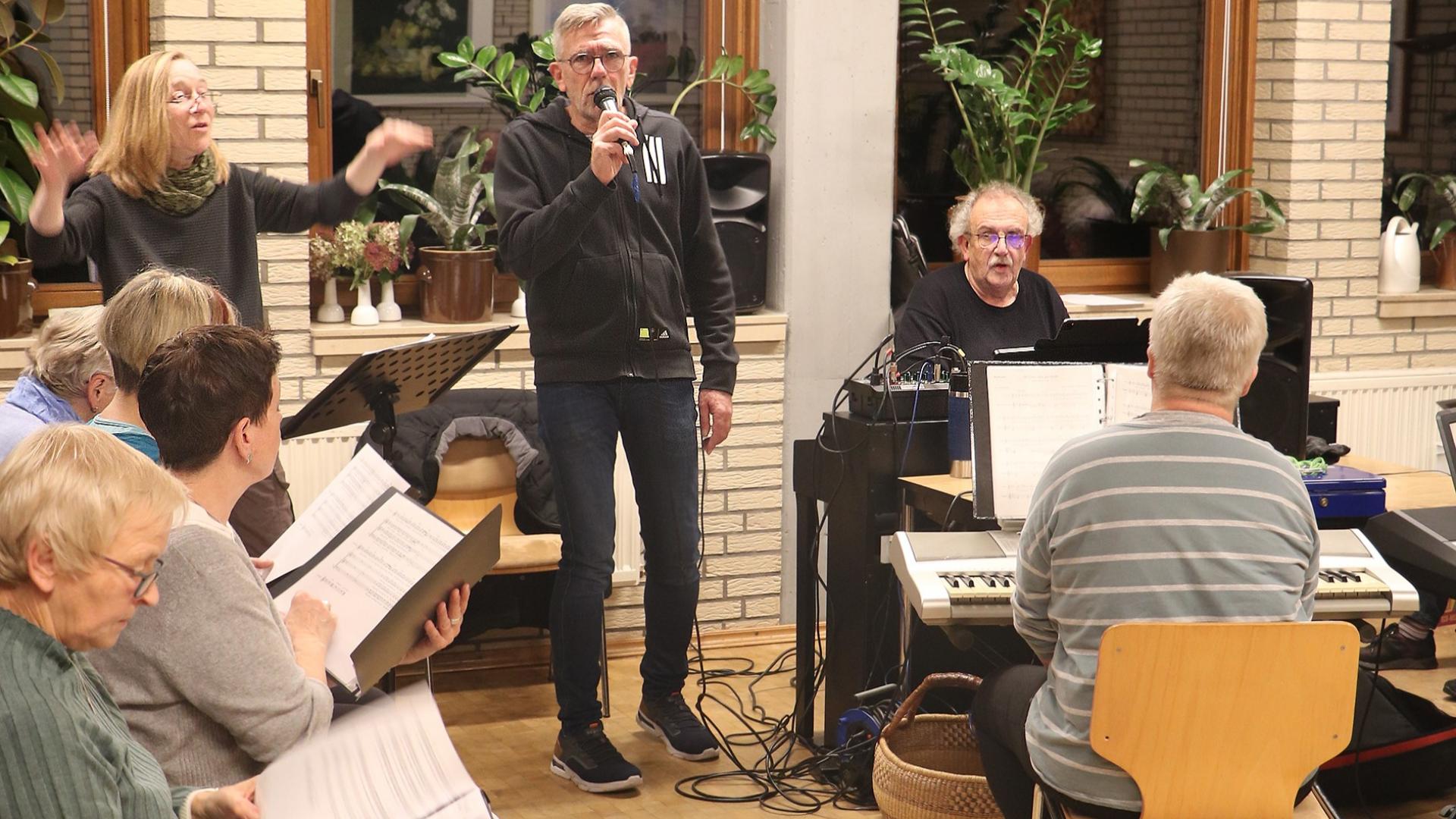 Ein Chor wird von einer Dirigentin geleitet. Ein Sänger singt sein Solo am Mikrofon. Begleitet werden die Sänger von zwei Personen am Klavier