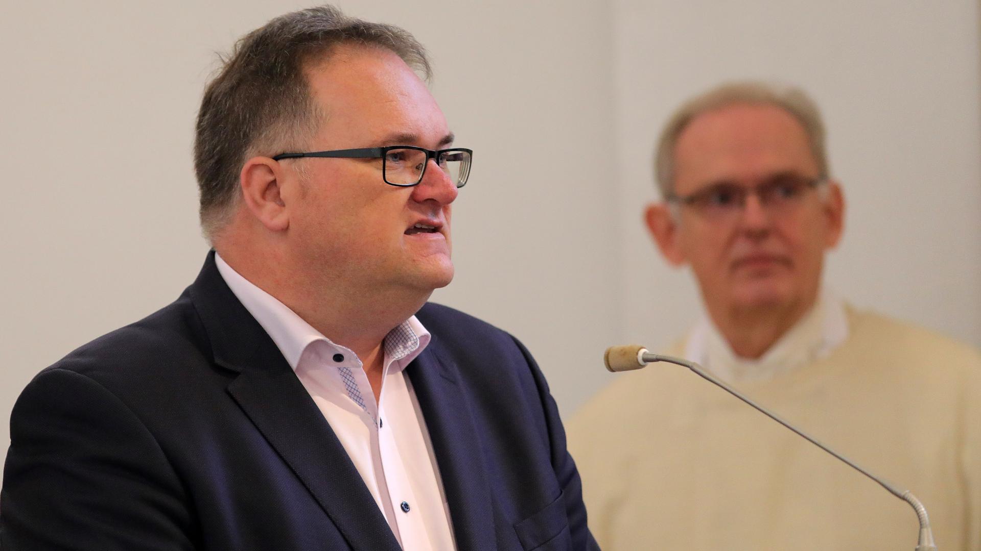 Ein Mann am Mikrophon, im Hintergrund ein Priester.