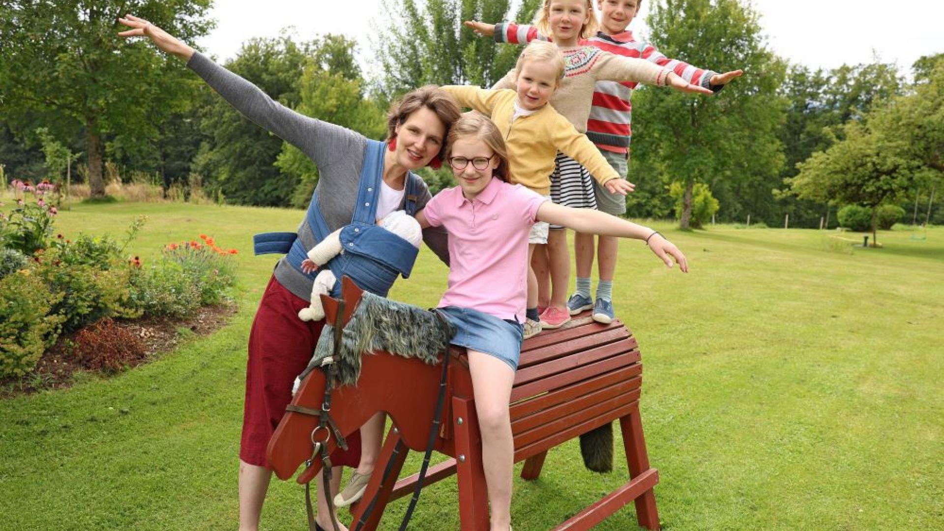 Vier Kinder spielen auf einem Holzpferd, ihre Mutter steht daneben und passt auf sie auf.