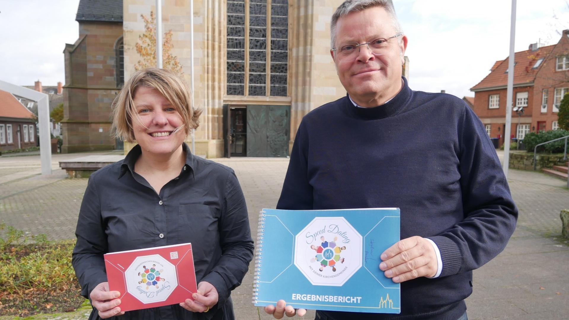 Eine Frau und ein Mann stehen auf dem Kirchplatz und halten zwei kleine Heftchen in die Kamera