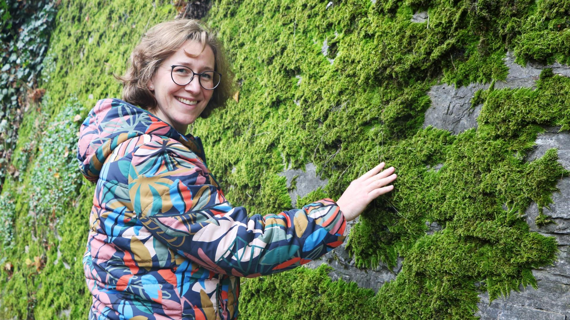 Eine lächelnde Frau führt ihre Hand zu einer moosbedeckten Wand