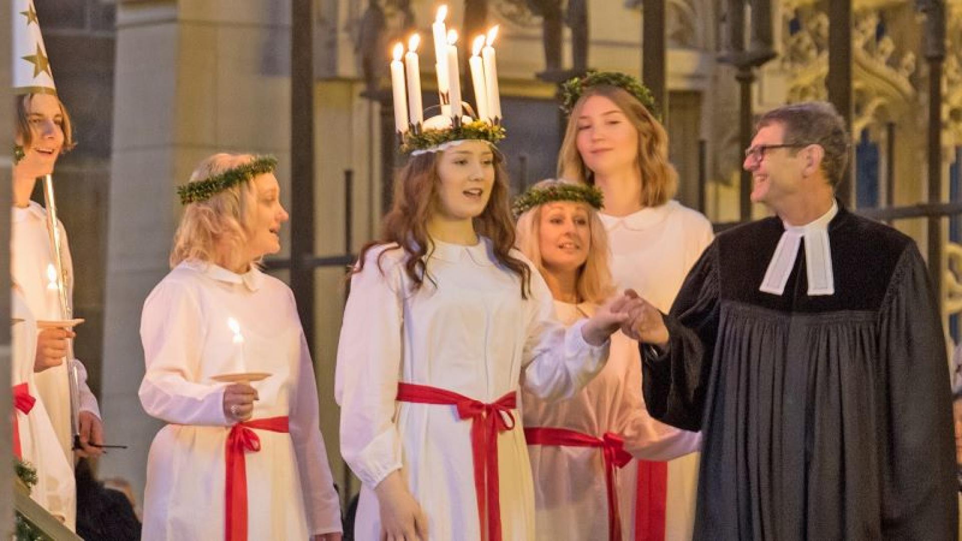 Eine junge Frau, die einen Kranz aus Kerzen trägt, wird von einem Pastor in die Kirche geleitet.