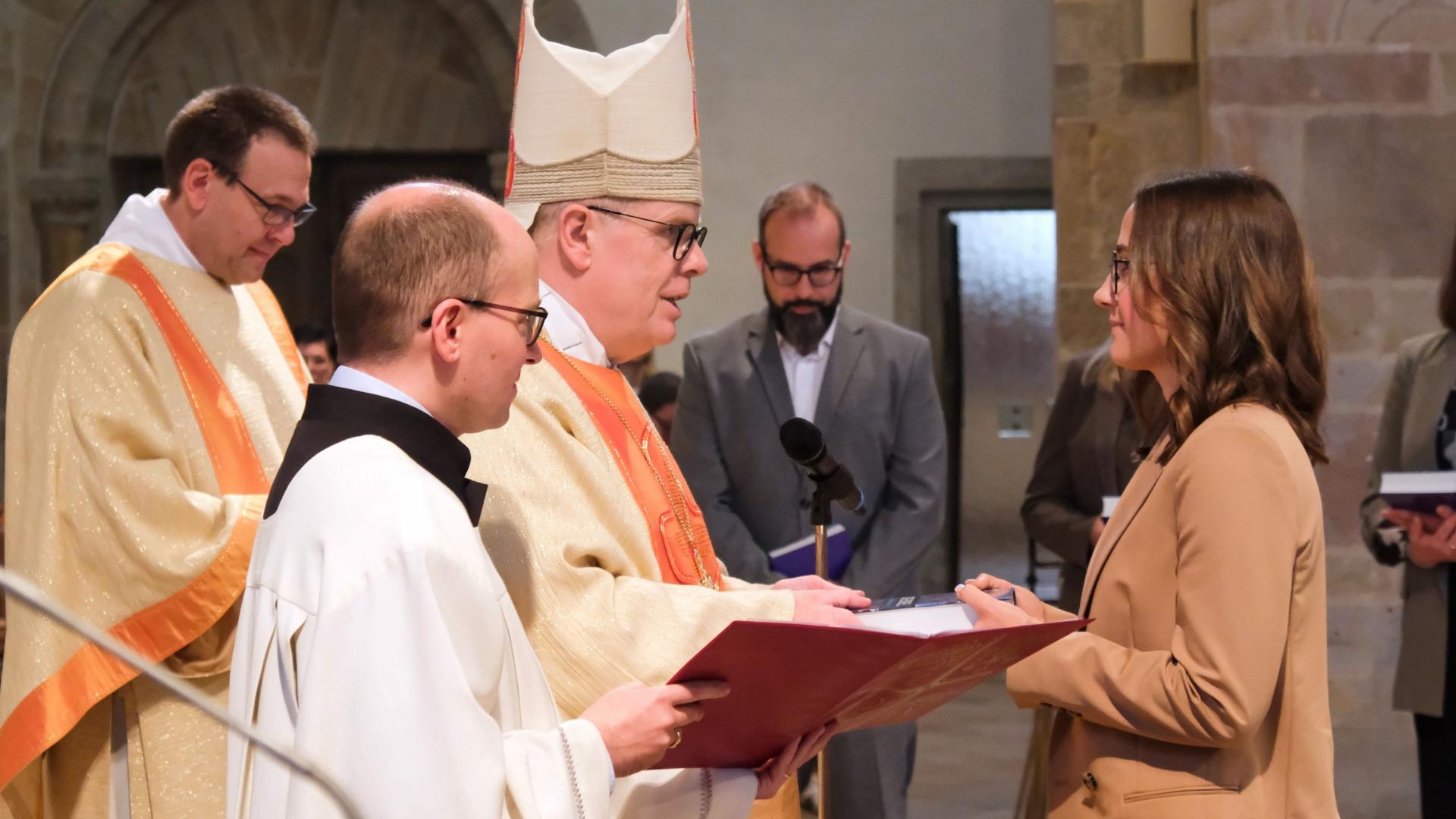 Ein Bischof mit Mitra auf dem Kopf steht vor einer jungen Frau. Neben ihm steht ein Ministrant mit geöffnetem Buch in der Hand