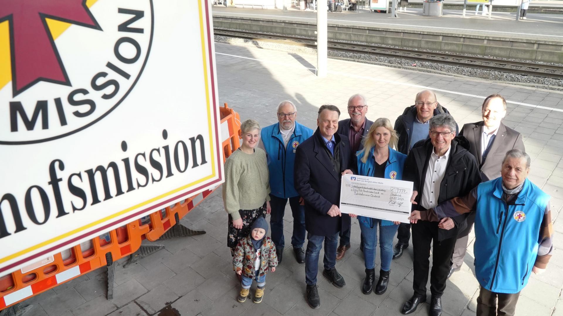 Mehrere Menschen stehen auf einem Bahnsteig und halten einen großen Scheck. Das Emblem der Bahnhofsmission ist zu sehen