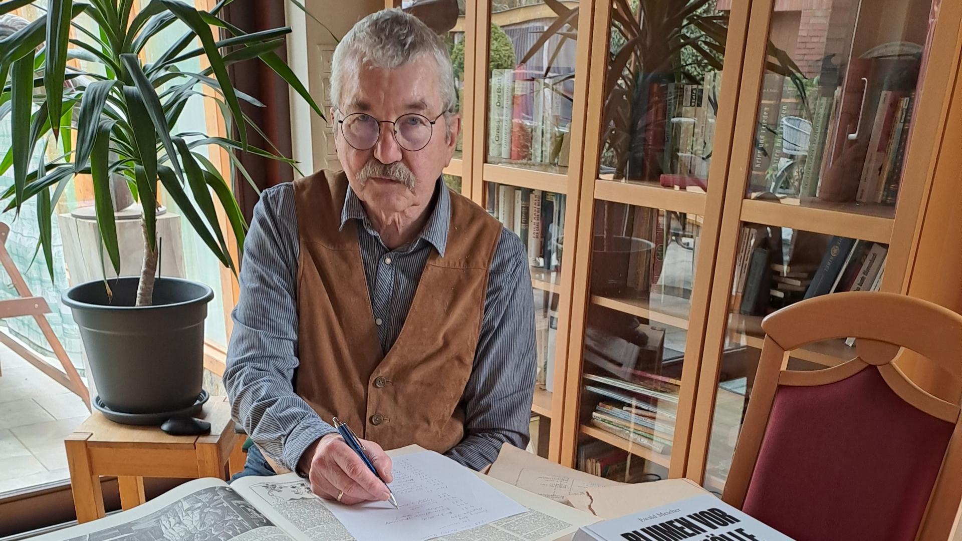 Ein älterer Mann sitzt an einem Tisch mit gestapelten Büchern und Blättern