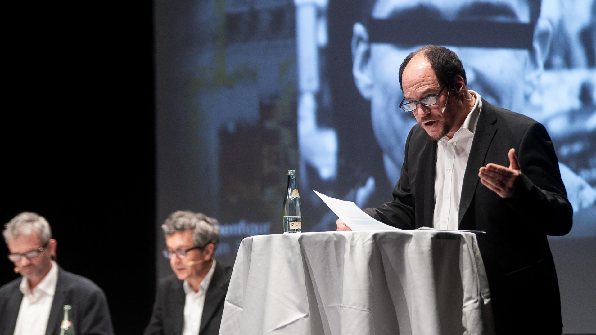 Ein Mann steht an einem Stehtisch und trägt einen Text vor.