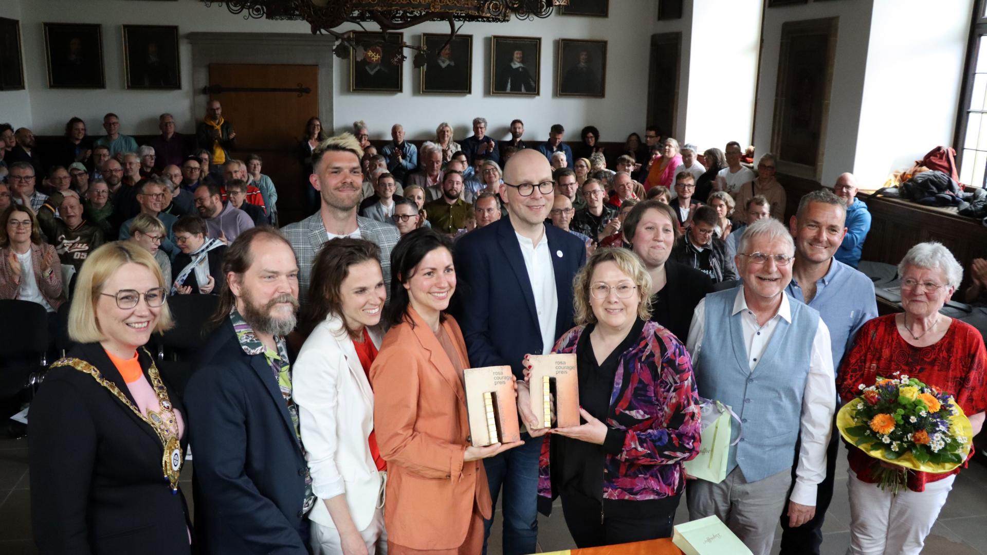 Mehrere Menschen stehen versammelt und halten die Preise in den Händen. Im Hintergrund sitzt ein Publikum