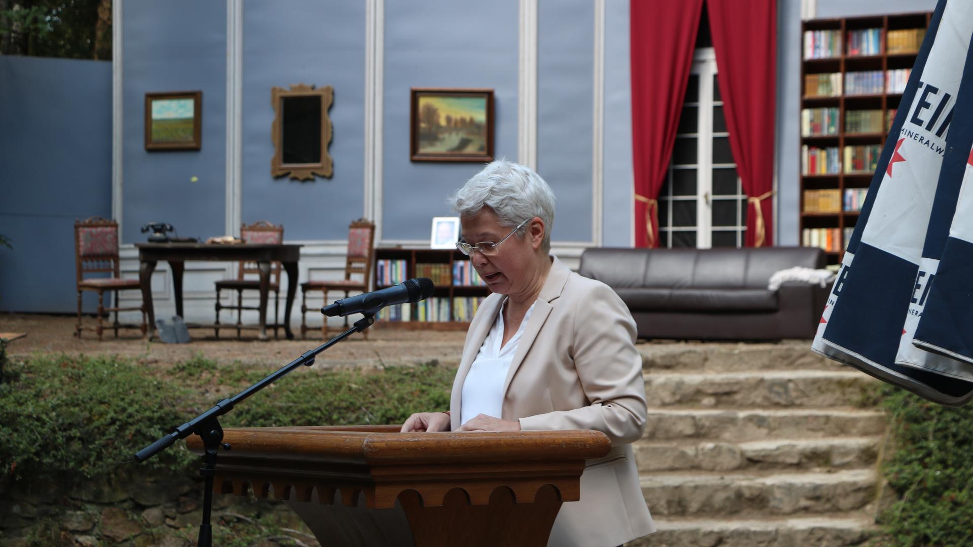 Frau an Stehpult mit Mikrofon vor einer Theaterkulisse.