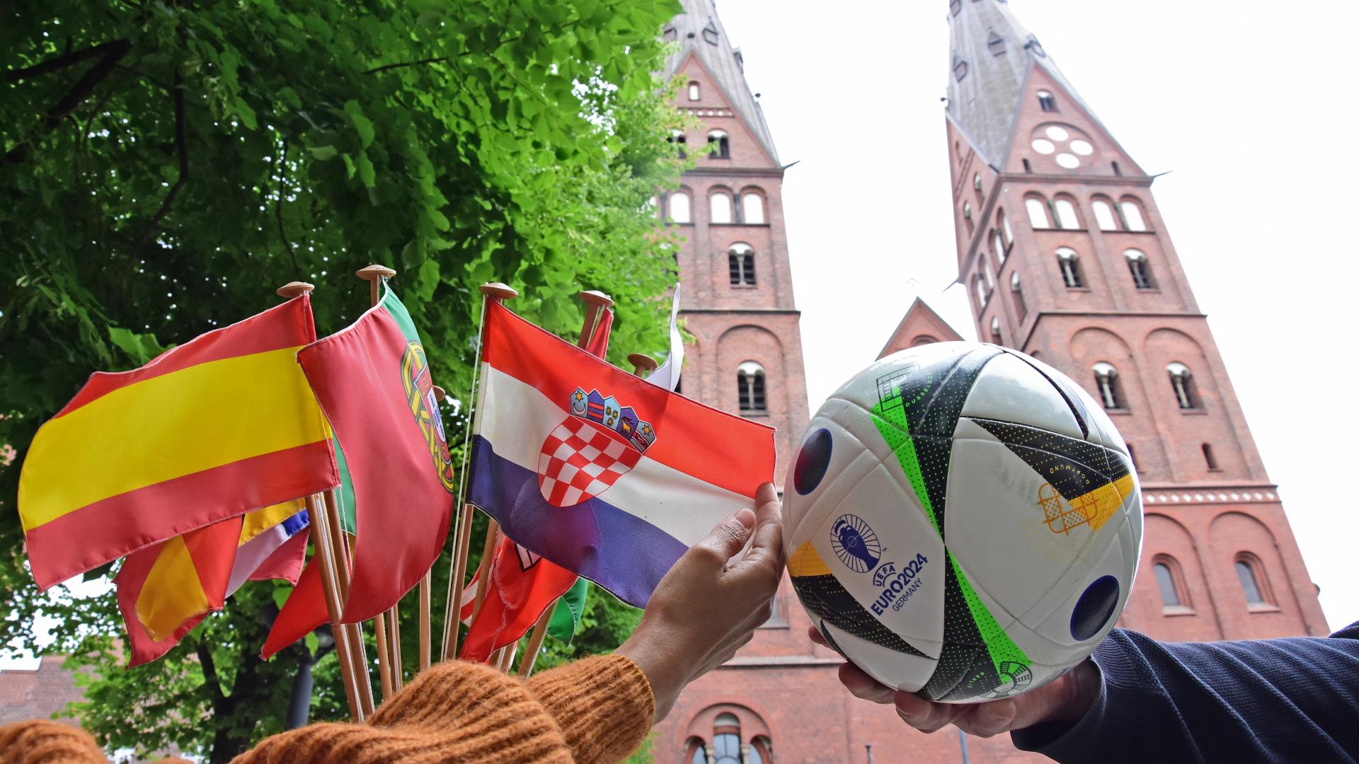 Hände zeigen Flaggen und einen Fußball vor dem St. Marien-Dom.