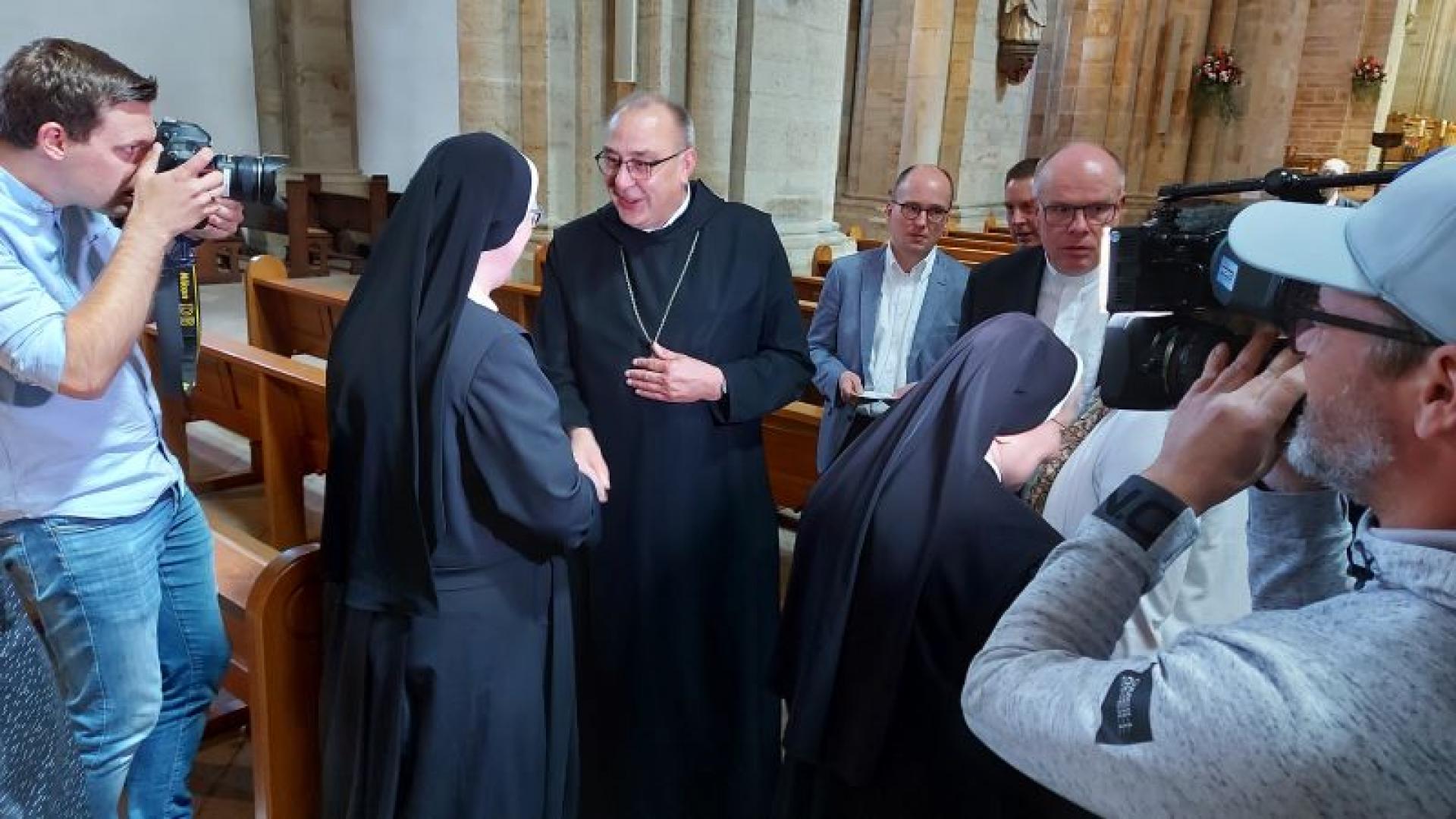 Eine Ordensschwester begrüßt den neuen Osnabrücker Bischof. Um sie herum stehen weitere Menschen sowie Kamerateams.