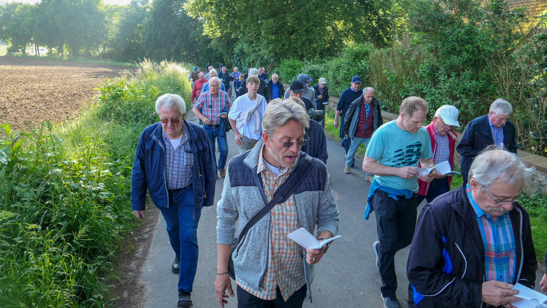 Eine Gruppe von Menschen geht einen Feldweg entlang. Einige haben Liederbücher in der Hand.